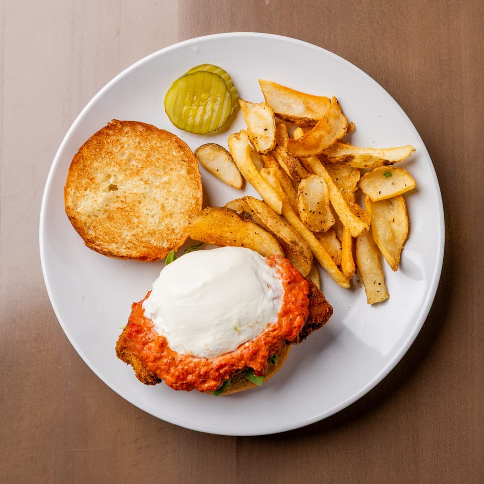 Chicken parmesan sandwich with melted mozzarella, red sauce, toasted bun, fries, and pickles on a white plate.