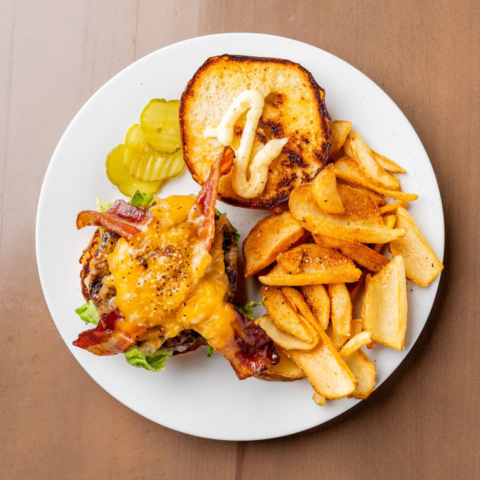 Overhead view of a bacon cheeseburger with fries and pickles on a white plate.