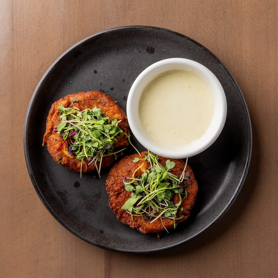 Two crispy fried crab cakes topped with microgreens, served with a side of creamy white sauce.