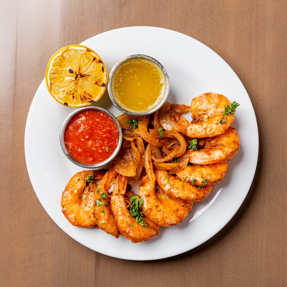 Grilled shrimp with sautéed onions, a charred lemon half, and two dipping sauces on a white plate.
